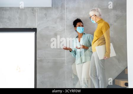 Female professionals in masks using laptop for technology, AI, digital innovation and safe workplace Stock Photo