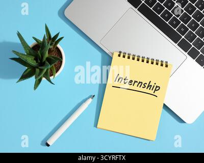 Yellow notepad with internship handwritten message, placed near laptop and pen on blue office desk Stock Photo