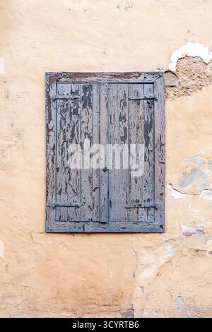 Peeling Blue Painted Wooden Window Shutter Set in an Old, Cracked Yellow Stucco Building Facade. Focus on Texture, Decay, and Vintage Charm Stock Photo