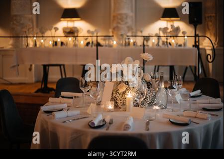 Elegant wedding reception table with white floral centerpiece, candles, and fine dining setup in a dimly lit room. Stock Photo
