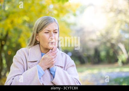 Senior woman clutching her cheek in an autumn park, grimacing from a sudden toothache and showing visible discomfort and distress while outdoors in casual fall clothing Stock Photo