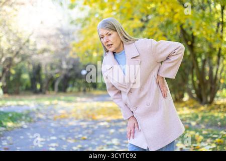 Senior woman clutching lower back in pain while walking through an autumn park, grimacing from chronic back ache and stiffness, posture bent from discomfort and mobility issues Stock Photo