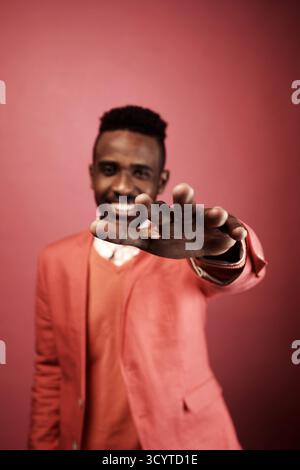 Portrait of defocused fashionable African American guy standing on pink background, stretching hand forward and smiling at camera cheerfully Stock Photo