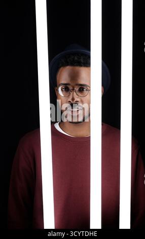 Black male fashionista with light sabers. Young stylish African American guy in stylish hat standing in dark studio behind fluorescent lamps and looking at camera Stock Photo