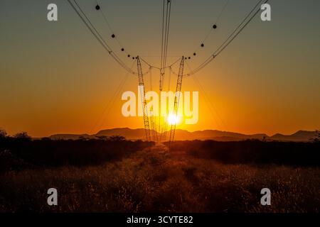 A line of power cables and pylons, with marker, vivibility balls, and the sun setting along the line, behind the Paresis Mountains in Namibia. Stock Photo