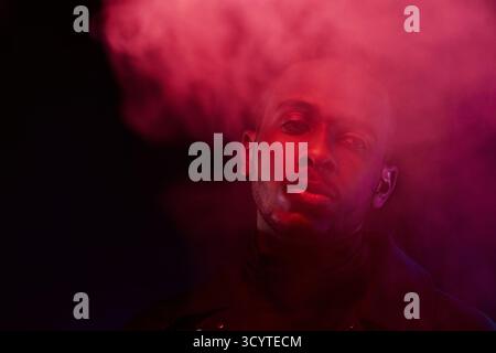 Portrait of young handsome Black man standing in smoke cloud illuminated by red light and looking away with tired face expression Stock Photo