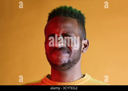 Portrait of young frustrated African American man with dyed green hair squinting his eyes in pain against yellow background Stock Photo