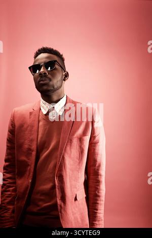 Studio portrait of young African American hipster man in stylish smart pink suit, shirt, knit jumper, sunglasses standing against plain red background, looking at camera Stock Photo