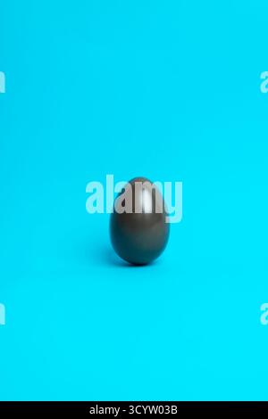 Single glossy chocolate Easter egg on bright blue background; minimalist festive concept for food photography, seasonal marketing, and holiday promos. Stock Photo