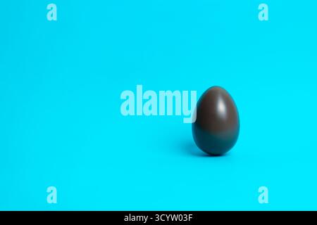 Smooth chocolate Easter egg standing on vibrant blue; minimal festive concept for holiday themes, confectionery branding, and seasonal promotions. Stock Photo