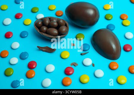 Whole and cracked chocolate Easter eggs filled with dark candies, surrounded by colorful candy-coated chocolates on vibrant blue. Stock Photo