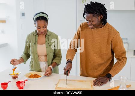 African American couple preparing layered pastry at counter using syrup bowl, red cups and knife Stock Photo