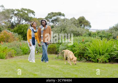 African American couple walking across garden lawn with yellow Labrador retriever sniffing grass Stock Photo