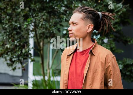 Man standing in courtyard wearing rust shirt under overshirt showing hair tie securing dreadlocks Stock Photo