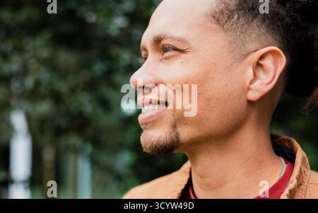 Mid adult man wearing red T-shirt, brown jacket standing in profile smiling at park, copy space Stock Photo