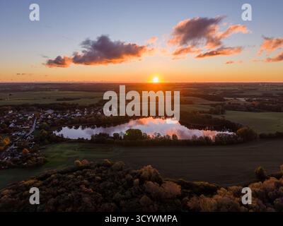 calm lake mirrors the vivid colors of a dramatic sunset sky, surrounded by open fields and a quiet village nestled in the peaceful countryside, Sildem Stock Photo