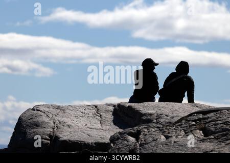 Two people sit on sunlit rocks under a bright blue sky in Tadoussac, Quebec, enjoying the peaceful coastal landscape. Stock Photo