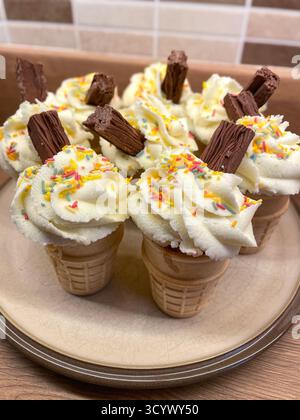 A plate of ice cream cone cupcakes topped with whipped cream, chocolate flakes, and colourful sprinkles Stock Photo