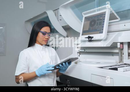 A woman wearing gloves and goggles examines data on a tablet in a clean, high-tech laboratory. Stock Photo