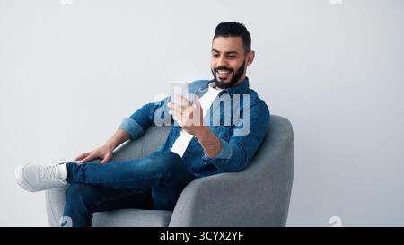Man Smiling While Using Smartphone in Modern Gray Chair Stock Photo