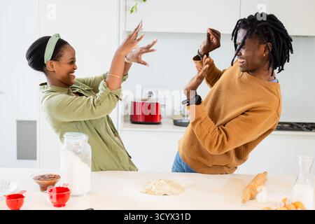 African American couple laughing and raising hands playfully in kitchen with flour jar, rolling pin Stock Photo