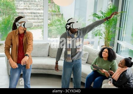 Diverse friends wearing VR headsets and exploring virtual world near sofa in bright living room Stock Photo