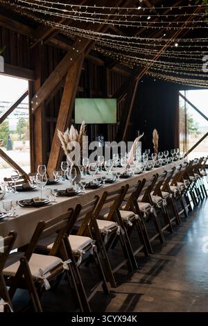 A long banquet table is elegantly set for a wedding reception inside a spacious rustic barn, featuring pampas grass centerpieces, warm string lights, Stock Photo
