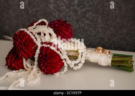 A close-up photograph features a lush bridal bouquet of deep red dahlias adorned with a delicate pearl necklace, showcasing gleaming gold wedding ring Stock Photo