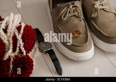 Romantic wedding morning scene showcasing gold rings atop brown suede shoes with a black watch and vibrant red floral bouquet. flatlay composition for Stock Photo