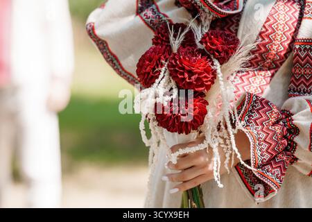 A close-up view showcases a bride's hands holding a vibrant bouquet of deep red dahlias and wheat, complemented by intricate embroidery on her white w Stock Photo