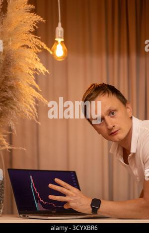 Thoughtful Young Man in Tense Focus Analyzing Cryptocurrency Market Charts and Trading Data on Laptop in Modern Home Office Workspace. Business, man. Stock Photo