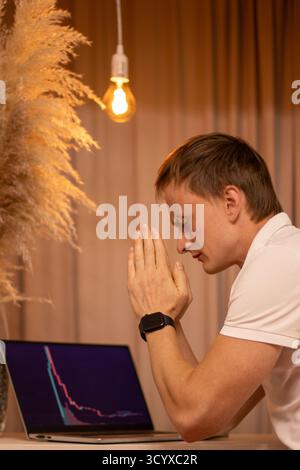 Thoughtful Young Man in Tense Focus Analyzing Cryptocurrency Market Charts and Trading Data on Laptop in Modern Home Office Workspace. Business, man. Stock Photo