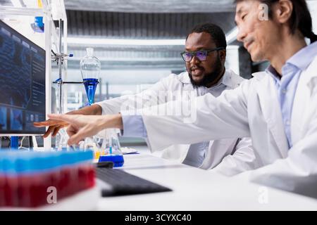 Team of biochemists in laboratory pointing to computer display, doing DNA data analysis. Research facility colleagues doing brainstorming using bioinformatics genomics software on PC in high tech lab Stock Photo