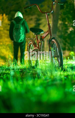 Backpacker Admiring Bicycle at Night. A person with a backpack stands contemplating a small red bicycle in a grassy area under night lights, foregroun Stock Photo