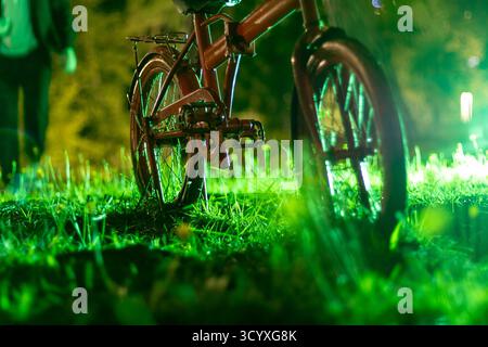 Side Close-Up of Bicycle in Grass. Horizontal close-up of a red bicycle's chain and rear wheel in green grass at night, with bokeh lights emphasizing Stock Photo