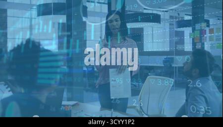 Presenting woman holding reports in modern office meeting, with laptop and stock data overlay Stock Photo