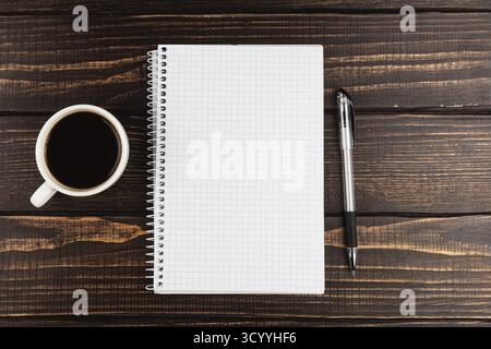 A blank notebook with a grid pattern sits next to a black coffee cup and a silver pen on a rustic wooden table. Perfect setup for note-taking or plann Stock Photo