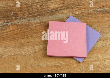 Two colorful sticky notes, one pink and one purple, rest on a wooden desk surface, ready for jotting down reminders or important tasks throughout the Stock Photo