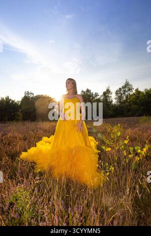 A woman elegantly poses in a stunning yellow gown amidst a blooming field as the sun sets beautifully Stock Photo