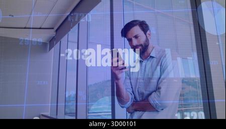 Standing businessman analyzing financial data overlays on glass wall in office, with smartphone Stock Photo
