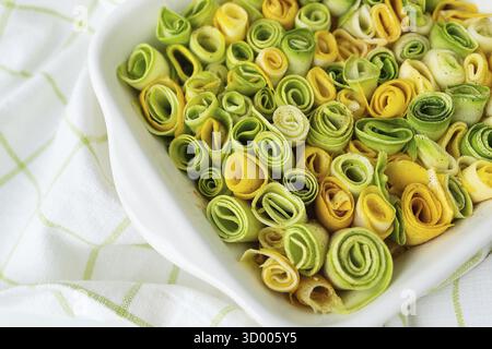 Small rolls of zucchini green and yellow with creamy sauce in a white baking dish against the background of a checkered napkin Stock Photo