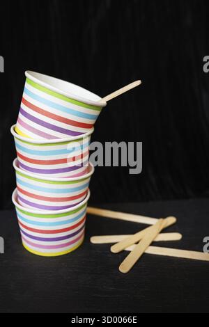 A stack of colorful striped paper cups and wooden stirrers on a black background. The cups are adorned with horizontal stripes in various bright color Stock Photo