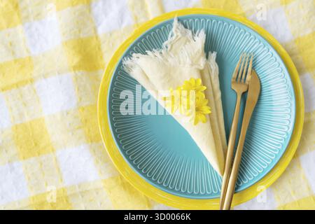 A bright blue plate with a yellow napkin, a wooden fork and fresh flowers on a checkered tablecloth. Table setting for the spring holiday Stock Photo