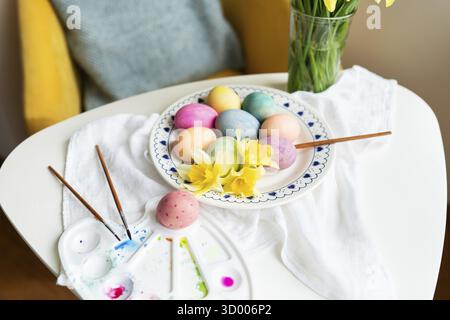 Pastel Easter eggs, daffodils, and paintbrushes are artfully arranged on a white table. This image evokes spring and the joyful celebration of Easter Stock Photo