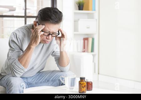 Portrait of 50s mature Asian man having headache, sitting on sofa at home, indoors living lifestyle Stock Photo