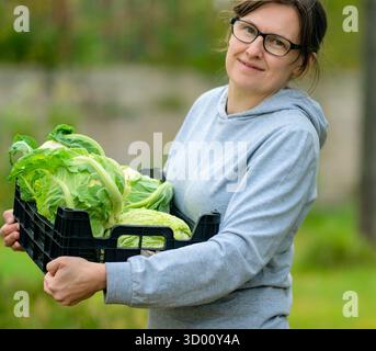 Smiling woman farmer holding a box full of fresh cauliflower during harvest season on her farm. Healthy vegetables grown naturally, organic produce rich in fiber and nutrients. Stock Photo