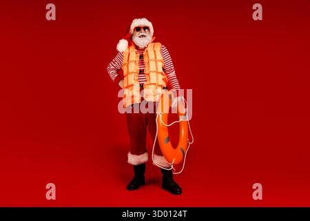 Santa in an orange life jacket holds a lifebuoy against a bold red studio background for a festive holiday rescue theme Stock Photo