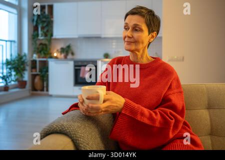 Calm mature woman drinking hot tea on sofa lost in thought in home comfort. Old female reflection Stock Photo
