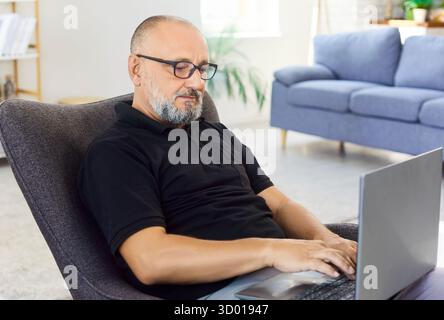 Senior man with glasses using laptop to work online or banking in living room Stock Photo