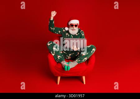 Santa in a festive pajama suit sits in a red chair on a bold red background holding a laptop with a raised fist celebrating Christmas cheer Stock Photo